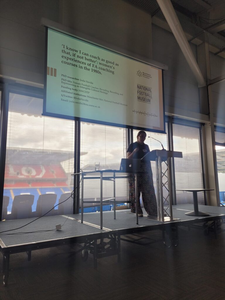 This photo shows Jodie Neville, speaking at the International Football History Conference at Cardiff City Stadium in 2024. I’m at the podium with a microphone, presenting in front of displayed slides. Stadium seating is visible through the adjacent window.